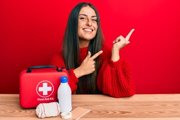 Beautiful hispanic woman with first aid kit smiling and looking at the camera pointing with two...