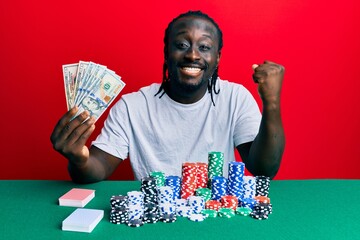Handsome young black man playing poker holding 100 dollars banknotes screaming proud, celebrating victory and success very excited with raised arm