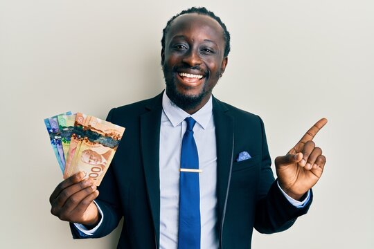Handsome young black man wearing business suit and tie holding canadian dollars smiling happy pointing with hand and finger to the side