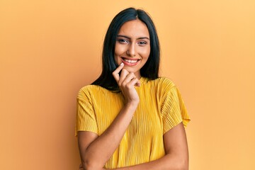 Young brunette woman wearing yellow tshirt over yellow background smiling looking confident at the camera with crossed arms and hand on chin. thinking positive.