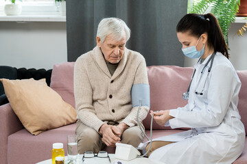 Young doctor in whitecoat and protective mask measuring blood pressure