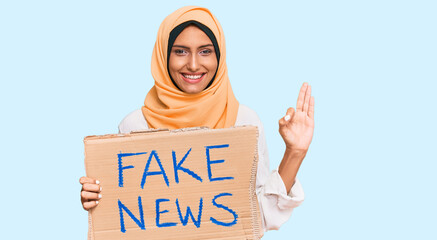 Young brunette arab woman holding fake news banner doing ok sign with fingers, smiling friendly...