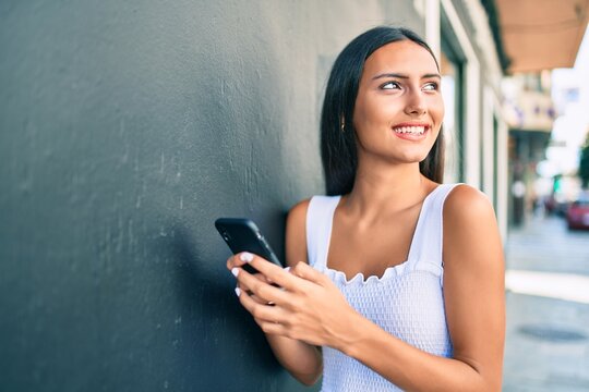 Young latin girl smiling happy using smartphone leaning on the wall.