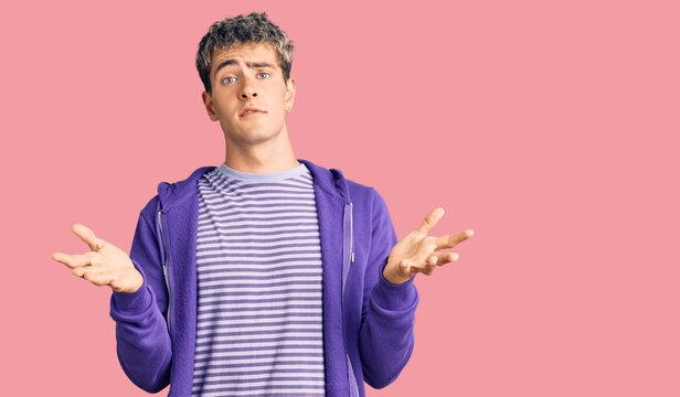 Young handsome man wearing casual purple sweatshirt clueless and confused expression with arms and hands raised. doubt concept.
