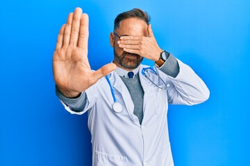 Handsome middle age man wearing doctor uniform and stethoscope covering eyes with hands and doing stop gesture with sad and fear expression. embarrassed and negative concept.