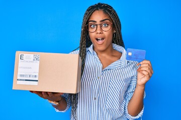 Young african american woman with braids holding delivery box and credit card afraid and shocked with surprise and amazed expression, fear and excited face.