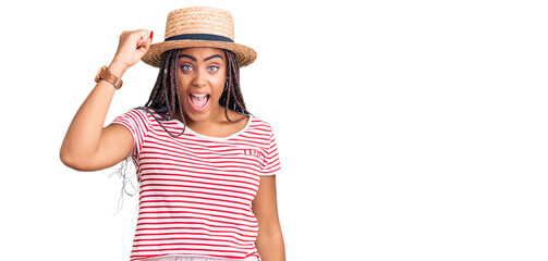 Young african american woman with braids wearing summer hat angry and mad raising fist frustrated and furious while shouting with anger. rage and aggressive concept.