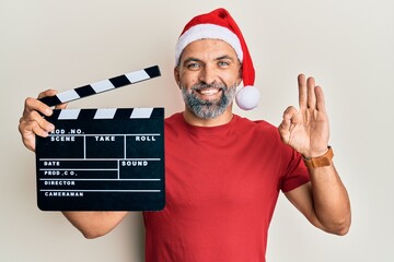 Middle age handsome man wearing christmas hat showing clapper doing ok sign with fingers, smiling...