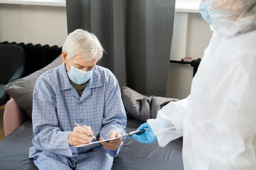 Sick senior male patient in mask and pajamas signing medical paper on couch