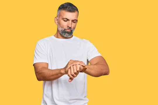 Middle age handsome man wearing casual white tshirt checking the time on wrist watch, relaxed and confident