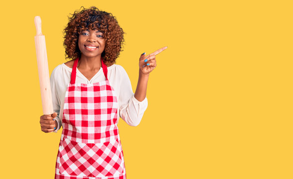 Young african american woman wearing apron holding rolling pin smiling happy pointing with hand and finger to the side