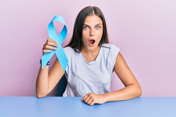 Young caucasian woman holding blue ribbon scared and amazed with open mouth for surprise, disbelief...