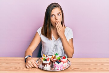 Young caucasian woman eating pastries for breakfast covering mouth with hand, shocked and afraid...