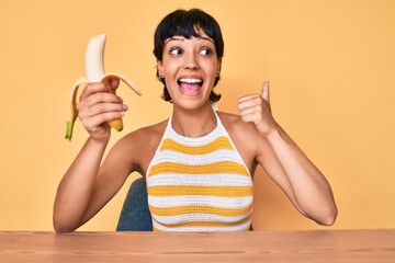 Brunette teenager girl eating banana as healthy snack pointing thumb up to the side smiling happy...