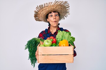 Beautiful brunettte woman wearing farmer clothes holding vegetables making fish face with mouth and squinting eyes, crazy and comical.