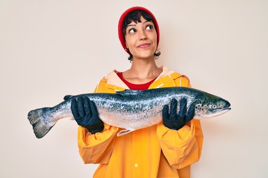 Beautiful brunettte fisher woman wearing raincoat holding fresh salmon smiling looking to the side and staring away thinking.