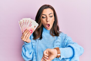 Young brunette woman holding 20 israel shekels banknotes looking at the watch time worried, afraid...