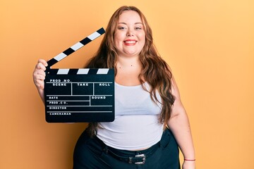Plus size caucasian young woman holding video film clapboard looking positive and happy standing...