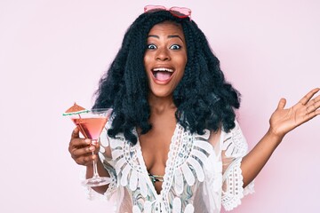 Beautiful african woman wearing summer look and drinking cocktail celebrating victory with happy smile and winner expression with raised hands