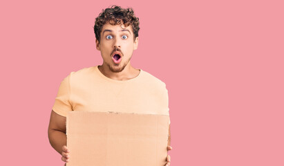 Young handsome man with curly hair holding blank cardboard empty banner scared and amazed with open...