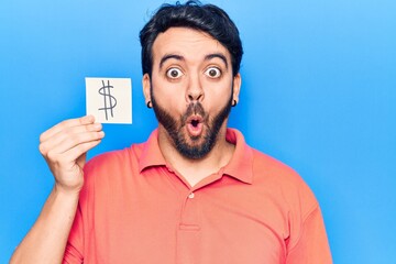 Young hispanic man holding dollar symbol reminder paper scared and amazed with open mouth for surprise, disbelief face