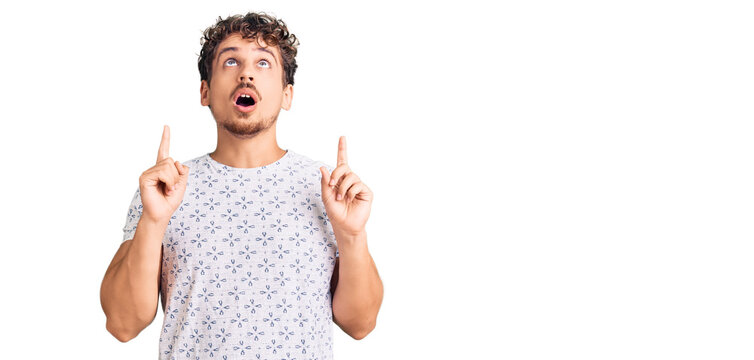 Young handsome man with curly hair wearing casual clothes amazed and surprised looking up and pointing with fingers and raised arms.