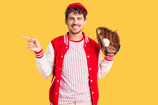 Young handsome man with curly hair wearing baseball uniform holding golve and ball smiling happy pointing with hand and finger to the side