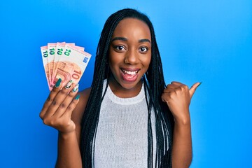 Young african american woman holding euro banknotes pointing thumb up to the side smiling happy...