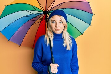 Young caucasian woman holding colorful umbrella relaxed with serious expression on face. simple and natural looking at the camera.