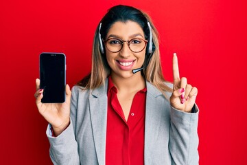 Naklejka premium Beautiful brunette woman wearing operator headset showing smartphone screen smiling with an idea or question pointing finger with happy face, number one