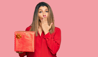 Beautiful brunette woman holding gift covering mouth with hand, shocked and afraid for mistake. surprised expression