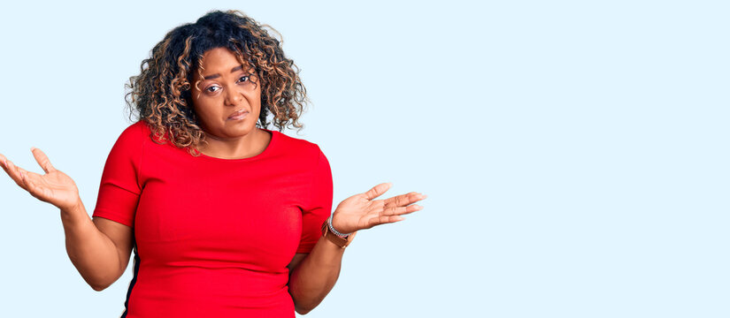 Young african american plus size woman wearing casual clothes clueless and confused expression with arms and hands raised. doubt concept.