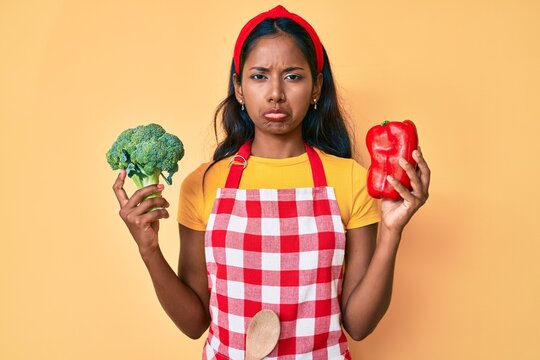 Young indian girl wearing apron holding broccoli and red pepper depressed and worry for distress, crying angry and afraid. sad expression.