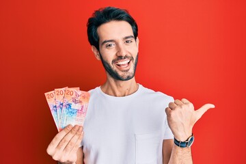 Young hispanic man holding swiss franc banknotes pointing thumb up to the side smiling happy with open mouth