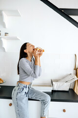 Portrait of rejoicing woman eats tasty croissant at home.
