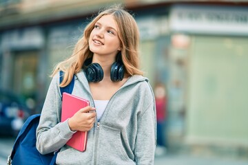 Beautiful caucasian student teenager using headphones holding book at the city.