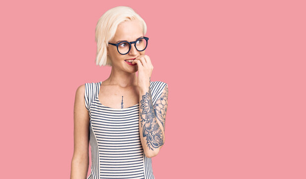 Young blonde woman with tattoo wearing casual clothes and glasses looking stressed and nervous with hands on mouth biting nails. anxiety problem.