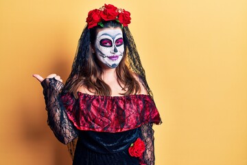 Young woman wearing day of the dead costume over yellow smiling with happy face looking and pointing to the side with thumb up.