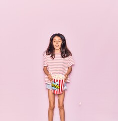Obraz premium Adorable hispanic child girl smiling happy. Jumping with smile on face holding bucket of popcorn over isolated pink background
