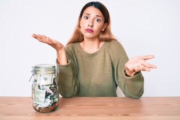 Young brunette woman sitting on the table with jar full of dollars clueless and confused expression...