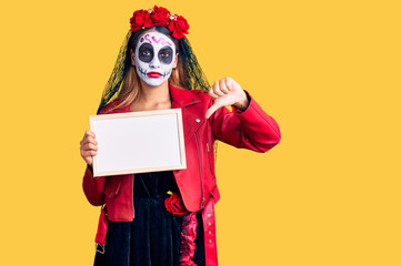 Woman wearing day of the dead costume holding empty white chalkboard with angry face, negative sign showing dislike with thumbs down, rejection concept