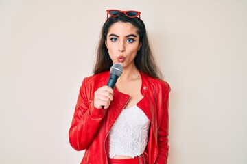 Brunette teenager girl singing song using microphone scared and amazed with open mouth for...