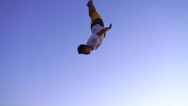 Young sports acrobat jumping on a trampoline. back flip. european guy in beachwear on blue sky background