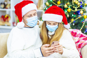 Couple in protective mask and santa claus hat talk online on phone. Boyfriend hold phone in her hand. Girlfriend wave hello to screen. Online meeting of Christmas amid the coronavirus pandemic