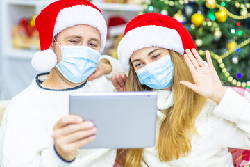 A young married couple with medical masks are sitting on the sofa in Santa hats against the background of a Christmas tree and congratulating relatives with Christmas (New Year) online using a tablet.