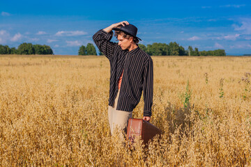 Tall handsome man with black hat and brown suitcase standing at golden oat field.