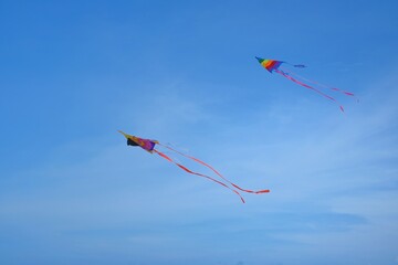 Two colorful triangle shape kites with rainbow pattern and long tail are flying parallel to each other against a clear blue sky and white fluffy clouds.