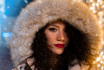 Portrait of beautiful young curly hair girl in winter furry jacket with hood, snow in background