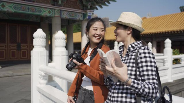 cheerful female temple visitors are leaning against the outdoor stone railing and pointing into distance while chatting with hearty laughs.