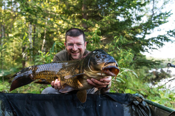 Happy smiling fisherman holding his fish  with lake on the background. Selective focus on the carp.